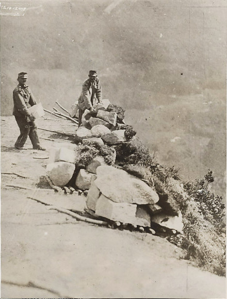 1915 photograph of Austro-Hungarian troops piling rocks to roll down upon Italian troops.
Text:
S/121415 Copyright by Underwood and Underwood.N.Y.
Austrians pile up huge stone boulders which they roll down the mountain side on Italian troops.
In their Alpine warfare on the Italians, the Austrians are resorting to a trick which is almost as old as the mountains themselves. They pile up huge boulders on the very edge of the mountain slope up which the Italians must make an ascent in order to attack the Austrian positions. The Austrians have but to use the eertion of raising the small crude cradle on which these boulders are resting, to start a veritable avalanche of stone, down the mountain side in the path of the on-coming Italians. The stone avalance is as effective as shot and shell for as it sweeps down the mountain side with the force of cannon fire, number of dead and maimed Italians are left in its wae. 9/23/15