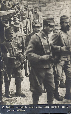 Cesare Battisti, in the background, indicated by a white cross over his head, being marched by Austro-Hungarian soldiers to his execution by garrotting, July 12, 1916. Battisti struggled for Italia irredenta, unredeemed Italy, Austro-Hungarian territory populated largely by ethnic Italians. The picture is from a series of photographs released by the Austrians as postcards.
Text:
C. Battisti scende le scale della fossa seguito dal Cappellano Militare.
C. Baptists descends the stairs of the ditch followed by a Military Chaplain.
Reverse:
Fotografia Depositata
Reproduzione Interdetta
Ed. Fot. S. Perdomi – Trento
Filed photograph; reproduction prohibited.